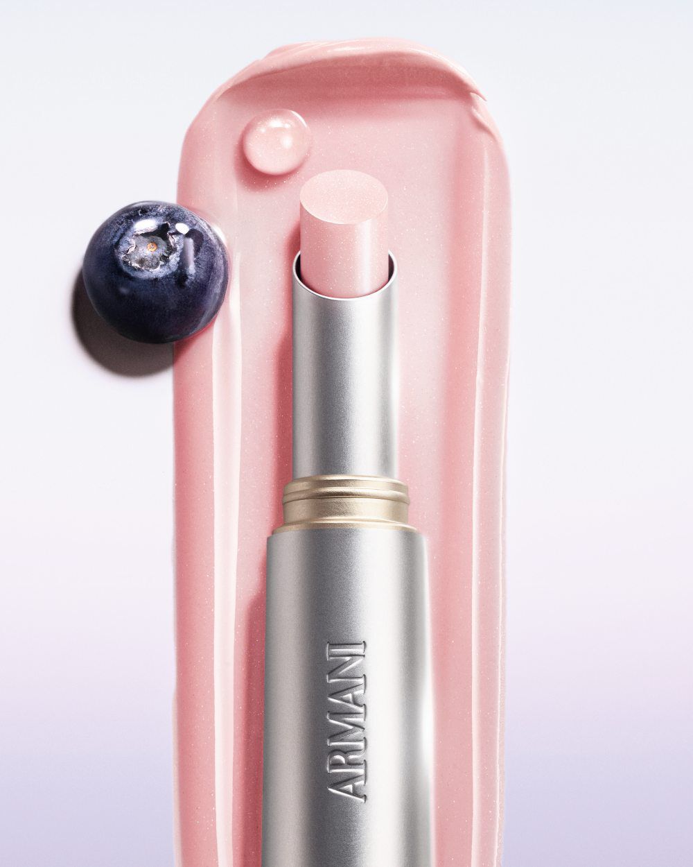 Armani Prisma Flash Petal Flash lipstick, captured in use, featuring its soft pink hue and elegant metallic packaging. Next to a blueberry fruit.