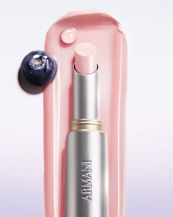 Armani Prisma Flash Petal Flash lipstick, captured in use, featuring its soft pink hue and elegant metallic packaging. Next to a blueberry fruit.