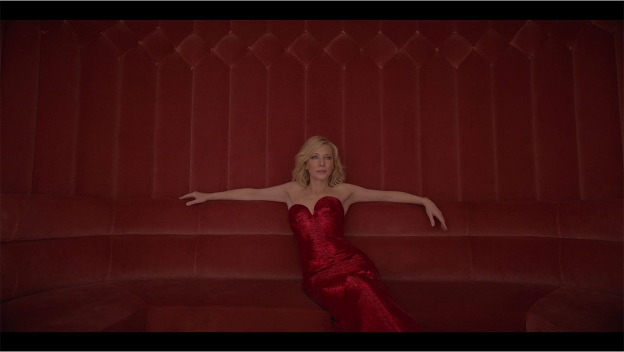 Woman with blonde hair wearing red strapless dress and arms winged out on red background
